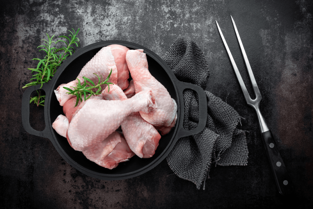 A black cast-iron skillet on a dark, textured background containing raw chicken drumsticks garnished with fresh rosemary, with a pair of carving forks to the side.