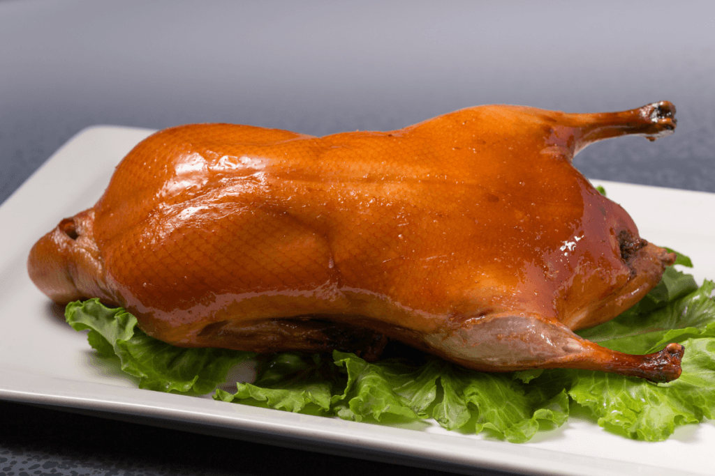 A roasted whole duck with crispy, golden-brown skin, presented on a white plate with a lettuce garnish.